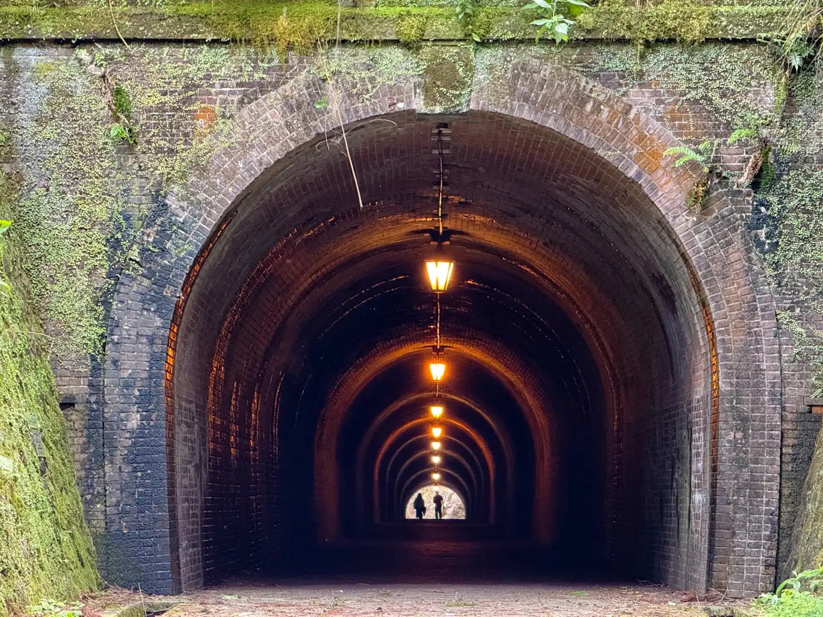 Standing at the tunnel mouth, looking straight through. Lamps hang down the brick vault, and two people are silhouetted at the far end.